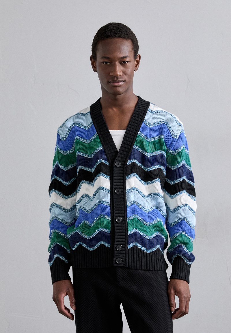 Multicolored knitted cardigan featuring a zigzag pattern in blue, green, black, and white. Ribbed black trim and buttons enhance the design.
