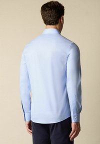 Lichtblauw langemouwenshirt met een klassieke kraag, gladde textuur en knoopmanchetten. Beschikt over zijsplitsen aan de zoom voor meer bewegingsvrijheid.