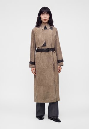 Personne avec de longs cheveux noirs portant un long manteau beige avec une ceinture noire et des détails, un pantalon large foncé et des chaussures noires pointues, debout.