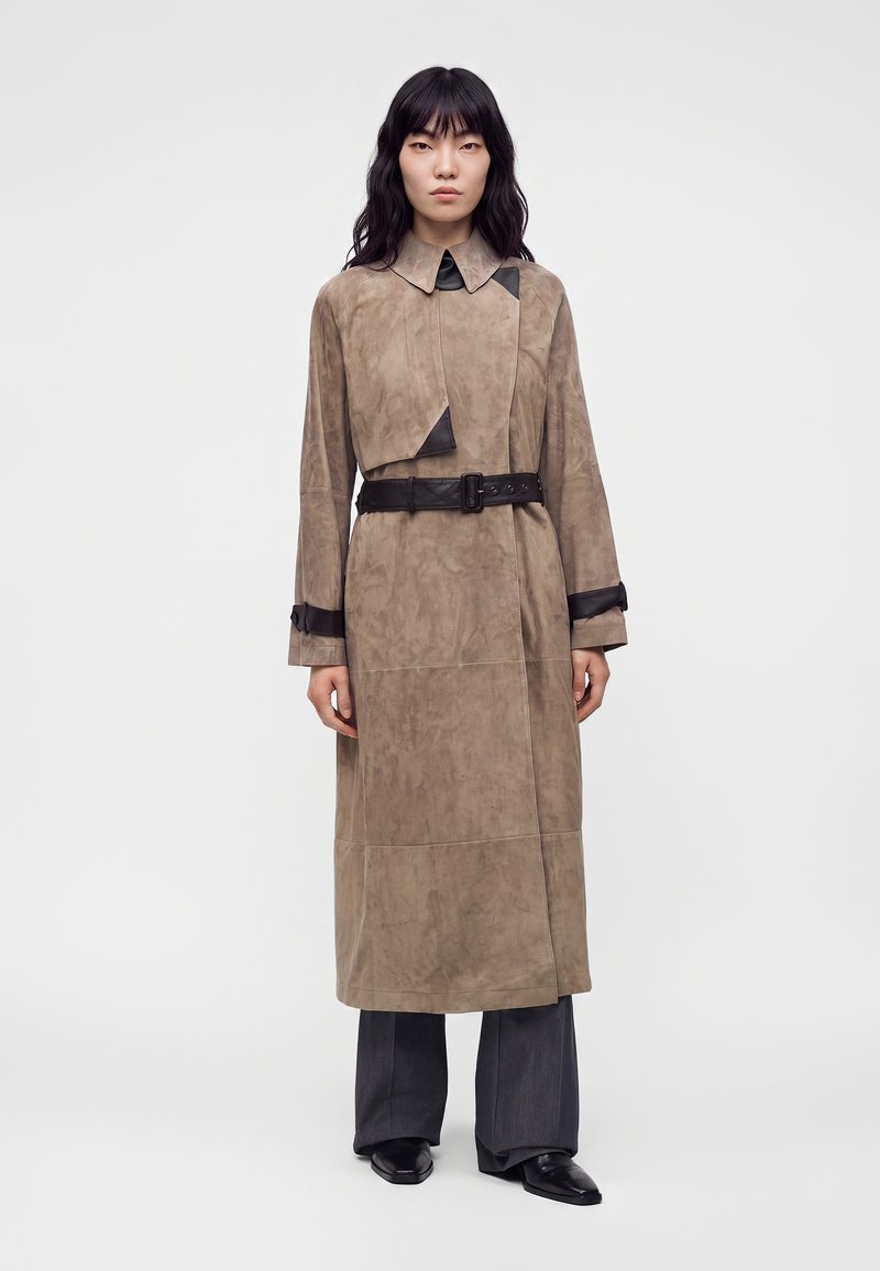 Personne avec de longs cheveux noirs portant un long manteau beige avec une ceinture noire et des détails, un pantalon large foncé et des chaussures noires pointues, debout.