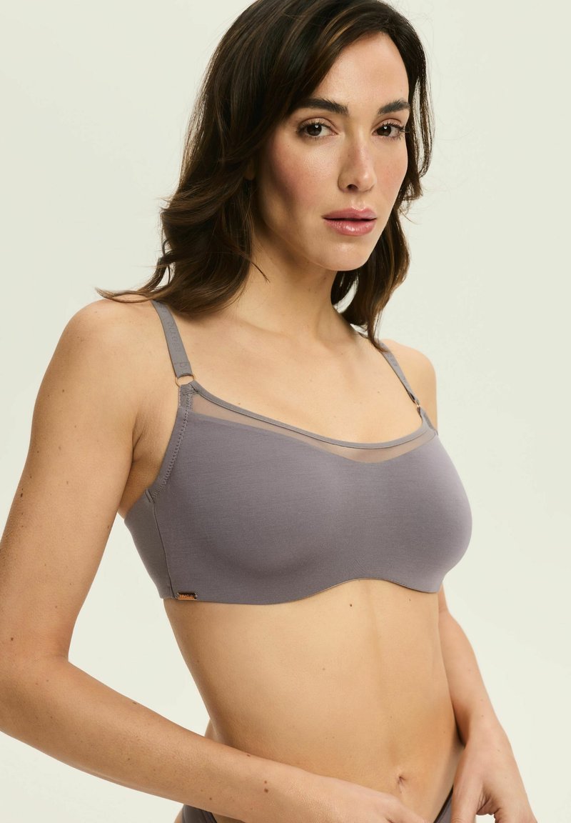 Szary biustonosz bezszwowy z regulowanymi ramiączkami, z przezroczystą górną taśmą i gładką teksturą, oferujący minimalistyczny design i wygodne dopasowanie.