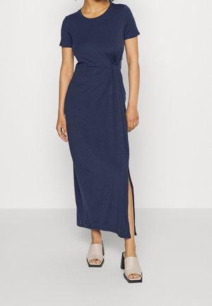 Femme portant une robe maxi bleu marine à manches courtes avec une fente latérale et des sandales beiges à talons blocs ouvertes à l’avant, debout sur un fond uni.