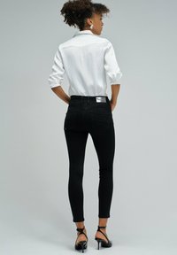Jeans skinny negros de cintura alta, con bolsillos traseros y una etiqueta de logo plateada; combinados con una camisa blanca con cuello y tacones negros con tiras.