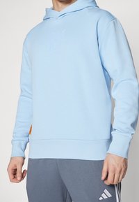 Persona che indossa una felpa con cappuccio blu chiaro con un logo sottile e pantaloni grigi Adidas con strisce bianche, in piedi con le mani rilassate.
