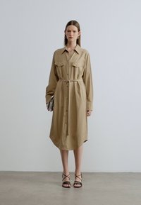 Καφές πουκάμισο-φόρεμα με κουμπιά, μακριά μανίκια, μπροστινές τσέπες, ζωνάκι στη μέση και μήκος μέχρι τη μέση της γάμπας, συνδυασμένο με μαύρα σανδάλια με λουράκια.