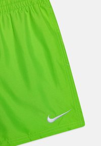 Limezaļa Nike šorti ar elastīgu jostu, uz kuriem netālu no apakšas ir redzams baltais Nike swoosh logo un pamanāmas šuvju detaļas.