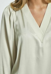 Blusa verde chiaro con scollatura a V profonda, morbida al tatto e leggermente plissettata sulle spalle. Maniche lunghe con una vestibilità comoda.