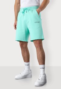 Shorts en coton turquoise avec une taille élastique, des poches latérales et des ourlets effilochés. Associés à des chaussettes blanches et des baskets avec logo.