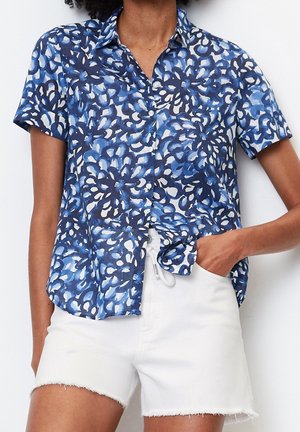 Kvinde iført en kortærmet skjorte med blå og hvide blomster og hvide denimshorts, hånd i lommen, mod en hvid baggrund.