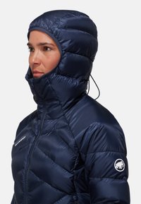 Marineblauwe geïsoleerde jas met een quilted ontwerp en een aansluitende capuchon. Voorzien van een ritssluiting en een logo patch op de linkerarm. Gladde, glanzende textuur.