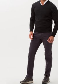 Uomo che indossa un maglione nero a girocollo, una camicia bianca con colletto, pantaloni grigio scuro e sneakers grigio scuro, in piedi contro uno sfondo bianco.