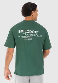 Camiseta de algodón verde oscuro con texto blanco en la parte posterior: "SMILODOX® TRAINING CLUB" con frases motivacionales adicionales debajo.