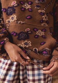 Brązowy, teksturowany hoodie z haftem w kwiaty w kolorze fioletowym i beżowym. Regulowany dół z brązowym troczkiem. W zestawieniu z wielokolorowymi spodniami w kratę.