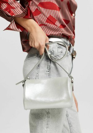 Bolso de mano metálico plateado con una textura suave, forma rectangular, correa ajustable y cierre de cremallera, sostenido en una mano contra unos jeans de mezclilla.