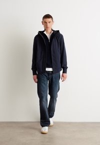 Marineblauwe zip-up hoodie, gecombineerd met een donkere trui, lichte blouse en donkere jeans. Witte sneakers met tan kleurige zolen. Minimalistisch ontwerp.