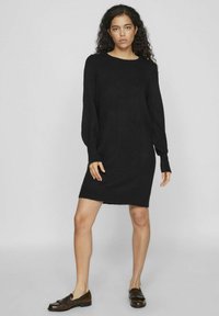 Abito nero in maglia con maniche lunghe, scollatura rotonda e una vestibilità leggermente ampia. Tessuto testurizzato con orlo sfrangiato. Indossato con mocassini marroni.