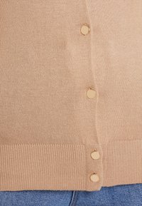 Jasnobeżowy kardigan wykonany z miękkiej dzianiny, z prążkowanym dołem i sześcioma okrągłymi, złotymi guzikami na listwie.