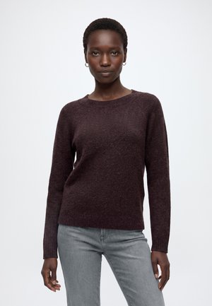 Femme aux cheveux courts portant un pull à manches longues marron foncé et un jean gris clair, debout devant un fond blanc uni.