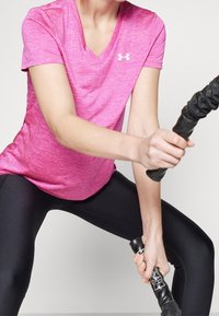 Rosa, v-ringad sporttopp med en melerad textur, kombinerad med svarta leggings. T-shirten har en liten logotyp på bröstet.