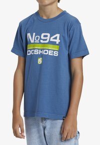 Modrá bavlněná tričko s krátkým rukávem, vyznačující se odvážným bílým a zeleným textem "No 94" a "DCSHOES", s volným střihem.