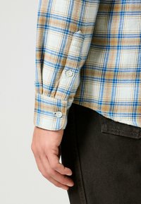 Geruite flanellen shirt in beige, blauw en wit. Lange mouwen met knoopmanchetten. Zachte textuur; gedragen met donkerbruine broek.