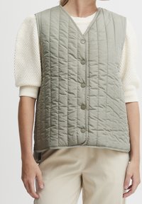 Gilet matelassé dans un vert doux et atténué, avec un col en V, des boutons sur le devant et une texture légèrement rembourrée. Porté par-dessus un pull en tricot.