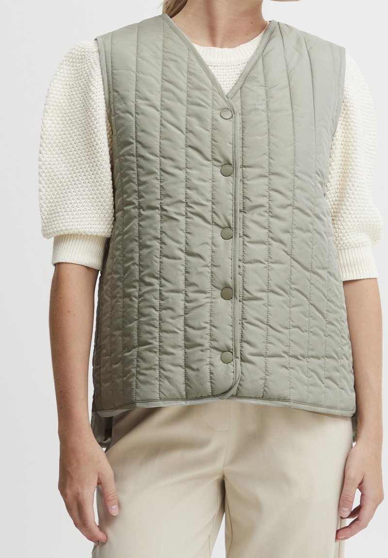 Gilet matelassé dans un vert doux et atténué, avec un col en V, des boutons sur le devant et une texture légèrement rembourrée. Porté par-dessus un pull en tricot.