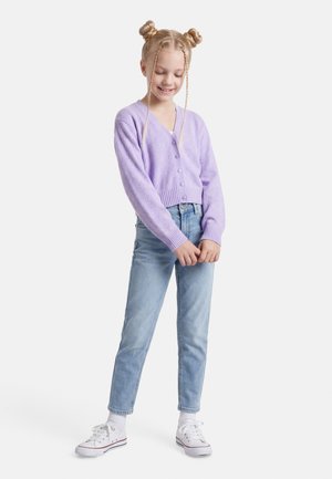 Fată tânără cu cocuri împletite, purtând un cardigan liliac cu nasturi, blugi albastru deschis, șosete albe și pantofi sport albi, stând în picioare și privind în jos.