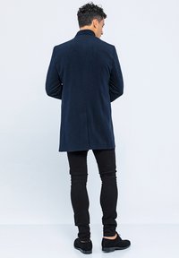 Marineblauwe wollen jas met een rechte snit, met een hoge kraag en een enkele split aan de achterkant, gecombineerd met zwarte slim-fit broeken en zwarte schoenen.