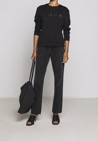 Schwarzer Sweatshirt mit geprägt "HUGO"-Logo, kombiniert mit dunklen Denim-Jeans und schwarzen, Riemchen-Sandaletten mit Absatz. Model hält eine schwarze Tote-Tasche.