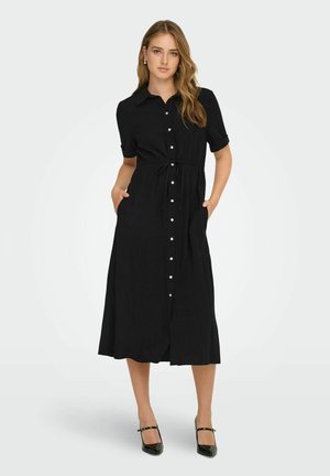 Kvinne som står med hendene i lommene iført en svart midikjole med knapper, korte ermer og svarte hæler mot en hvit bakgrunn.