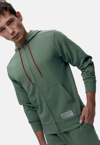 Kapuzensweatshirt mit Reißverschluss aus olivgrünem Stoff, mit roten Kordeln, einer Kängurutasche und einem Etikett auf der unteren linken Seite.