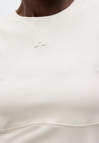 Κοντινό πλάνο ενός ατόμου που φοράει λευκή ριμπ μπλούζα με ένα μικρό, ασημένιο, αφηρημένο λογότυπο στο κέντρο κοντά στην κλείδα.