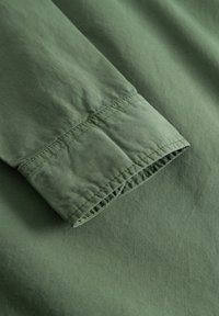 Tessuto di cotone verde con una texture liscia, caratterizzato da un polsino con dettagli di cucitura discreti. In generale, un design semplice e funzionale.