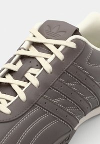Brown leather sneaker featuring white laces, zigzag stitching accents, and perforations along the sides. Brand logo embossed on tongue.