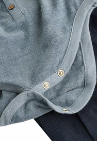 Helles blaues Kurzarmoberteil mit Knopfkragen und metallic-farbenen Druckknöpfen, über einem dunklen gemusterten Stoff geschichtet, der eine glatte Textur aufweist.