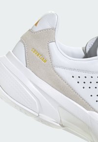 Baskets blanches en cuir et daim avec un design perforé, un col rembourré et un détail doré "COURT" sur le côté. Semelle extérieure texturée blanche.