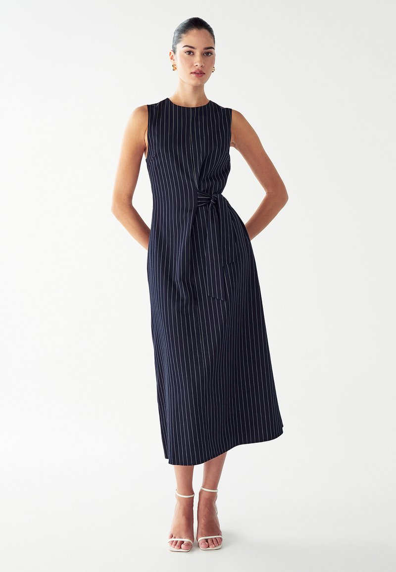 Mouwloze marineblauwe jurk met witte pinstrepen, geknoopte taille-informatie en een midi-lengte. Glad materiaal met een ontspannen silhouet.