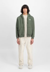 Sweatshirt de algodão verde com cordões, dois bolsos frontais e detalhe de logótipo, combinado com calças cargo creme e sapatos claros.