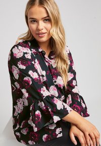 Blusa floreale con sfondo nero, caratterizzata da fiori rosa e bianchi, con bottoni frontali, colletto e maniche svasate. Tessuto morbido.