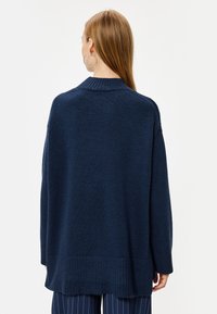 Maglione lavorato a maglia blu navy con collo alto, spalle scese e bordo a costine. Texture morbida e vestibilità rilassata, indossato sopra pantaloni a righe.