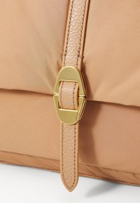 Zbliżenie na beżową torbę z tkaniny, z teksturowanym uchwytem i złotą klamrą, podkreślającą gładkie wykończenie i miękki wygląd.