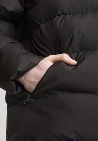 Juodas pūkinis švarkas su tekstūruotu paviršiumi, siuvinėjimu ir šonine kišenė su užtrauktuku. Reguliuojama medžiaga užtikrina šilumą ir komfortą.