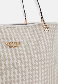 Sac fourre-tout tissé beige avec des accents blancs, présentant des accessoires en or et un logo. Le sac dispose de deux anses noires et a une forme structurée.