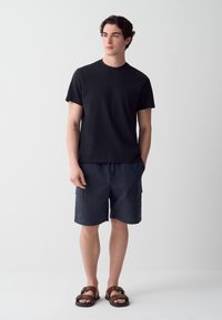 T-shirt nera a maniche corte, pantaloni cargo blu navy con tasche e sandali marroni con cinturino; outfit casual con vestibilità rilassata.