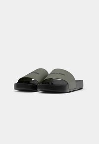 Melnas un tumši zaļas Calvin Klein čības ar izceltām zoles uz baltas fona.