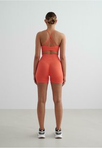 Oranžová športová podprsenka s prekrývajúcimi sa ramienkami a zodpovedajúcimi šortkami s vysokým pásom. Materiál sa zdá byť elastický. Model má na sebe biele športové topánky.