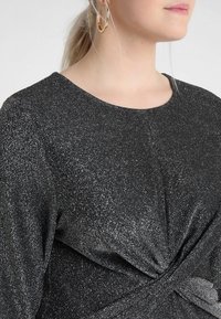Černý top s kulatým výstřihem, vyrobený z texturované, třpytivé tkaniny. Detail uzlu v pase zvýrazňuje design.