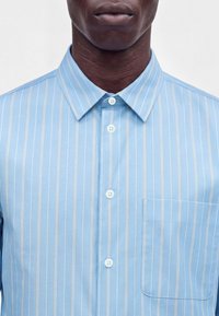 Homme portant une chemise bleu clair à fines rayures verticales blanches, boutonnée avec un col et une poche poitrine sur le côté droit.