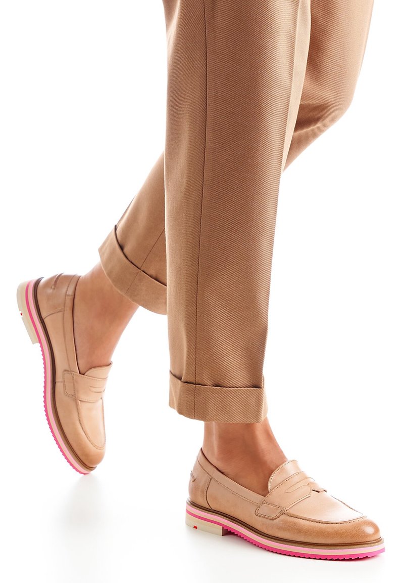 Personne portant des mocassins beige à semelles roses et un pantalon brun retroussé, marchant sur un fond blanc.
